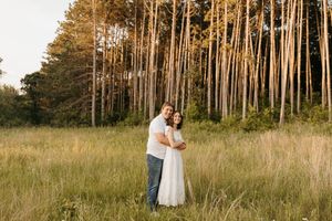 Couple embracing in tall grass field with pine forest backdrop during golden hour sunset.