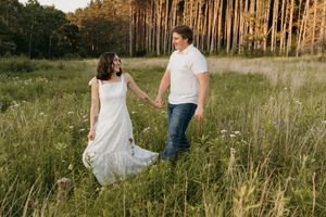 Couple holding hands walking through wildflower meadow near pine forest during golden hour.