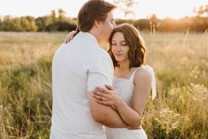 Couple embracing tenderly in golden sunlit meadow, woman in white dress with eyes closed.