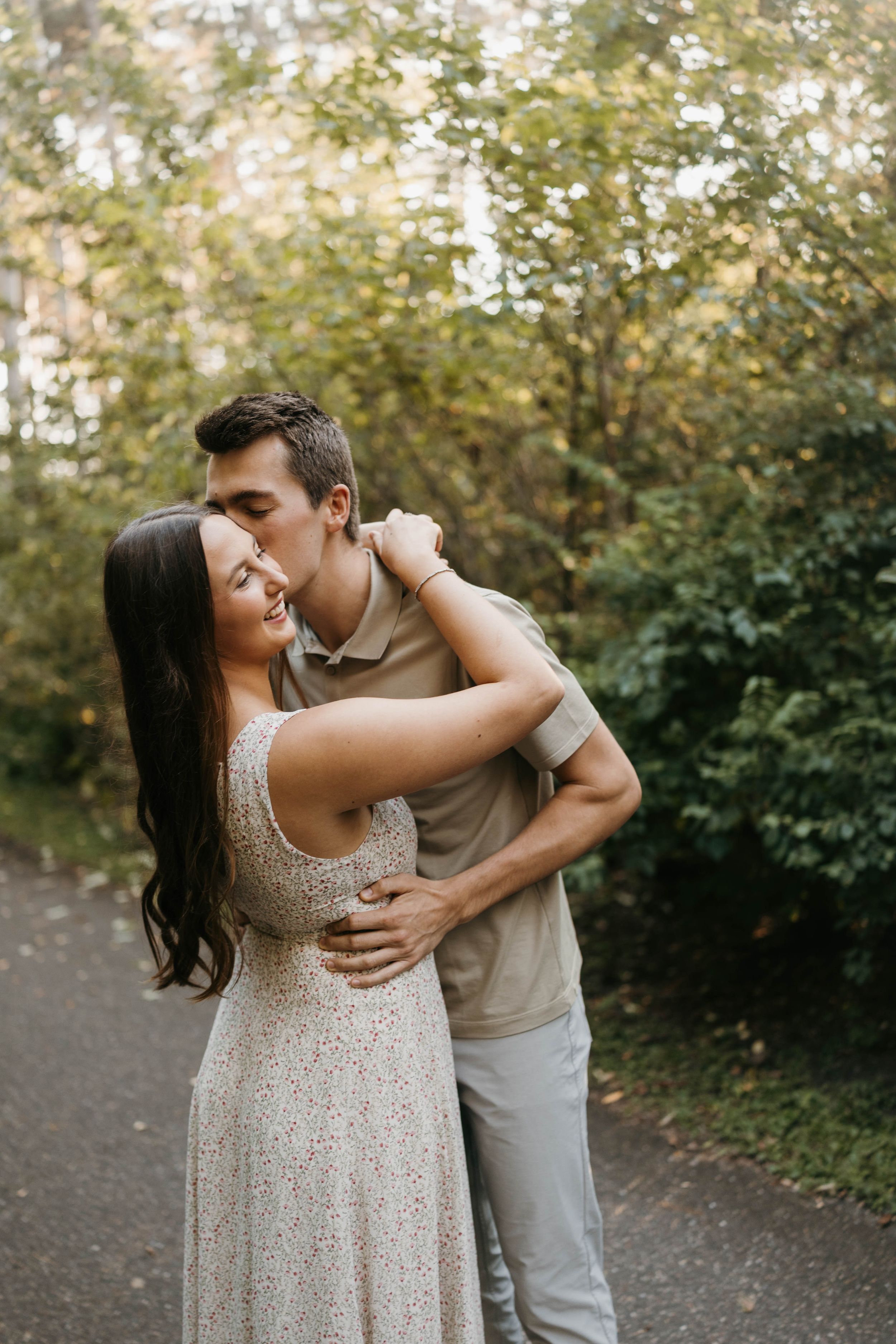 Man kisses woman on the cheek as she smiles during a romantic outdoor engagement session among green trees.