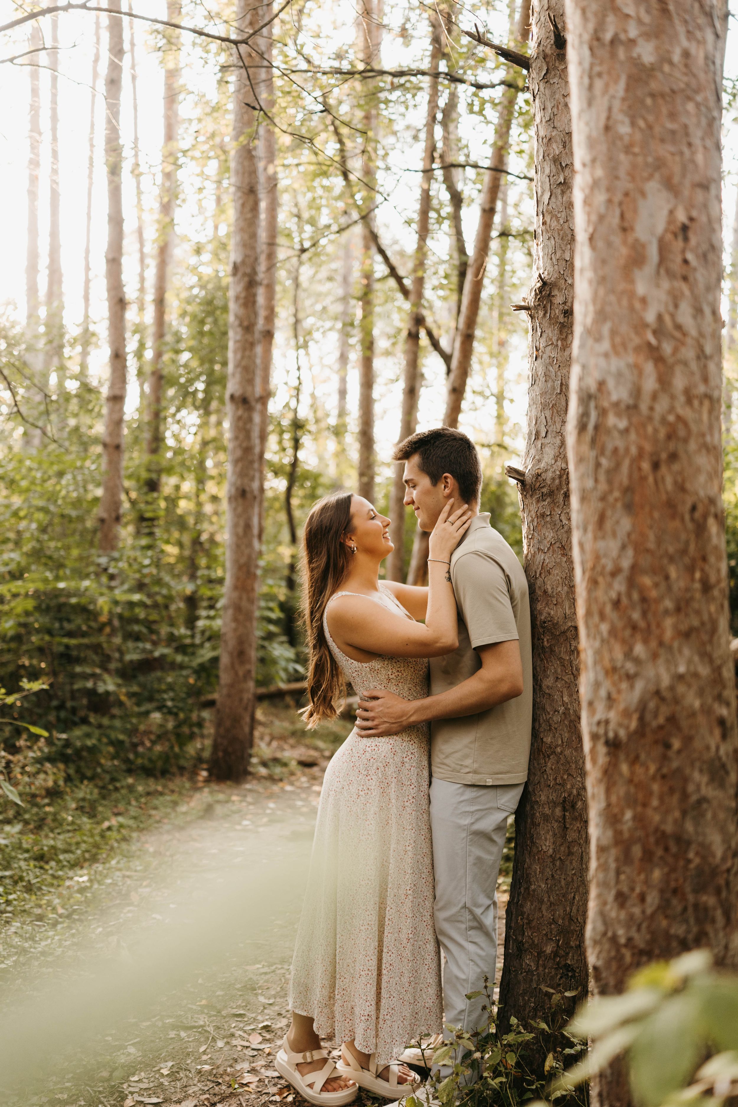 Couple embracing romantically against a tree in a sunlit forest, woman in floral dress gazing up at man.