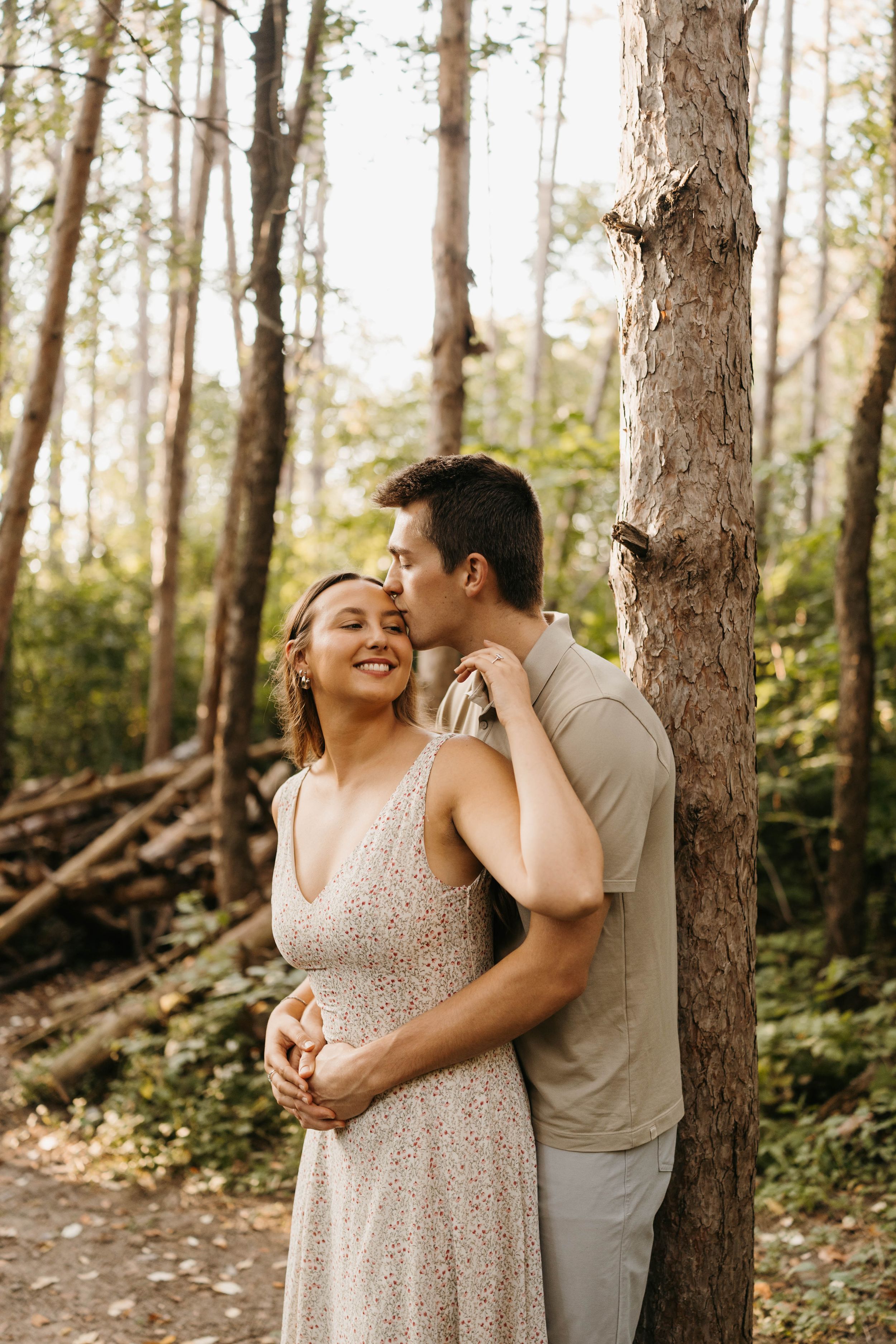 Man kissing woman on cheek while embracing her from behind near a tree in a lush green forest.