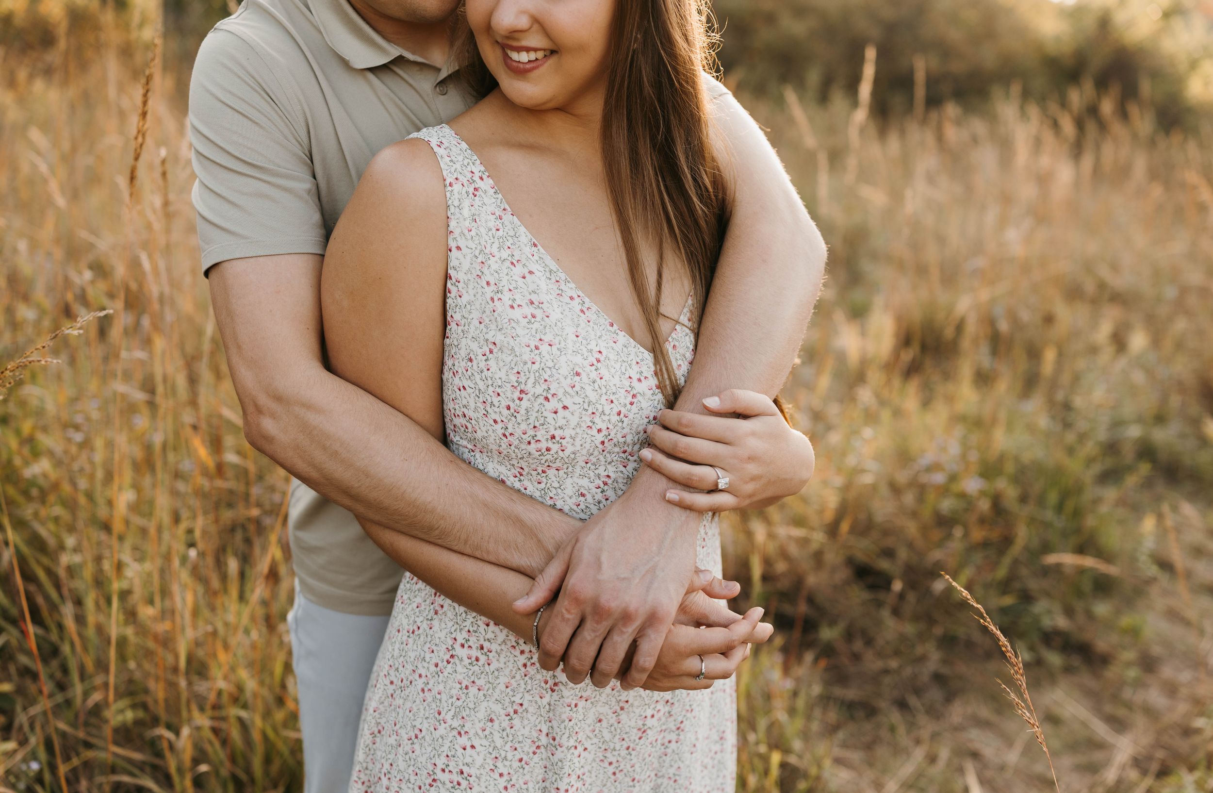 Close-up of couple embracing in golden field, woman in floral dress wearing engagement ring.