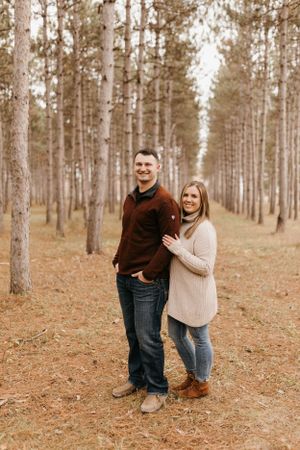 Couple posing in tall pine forest, man in burgundy sweater and woman in beige sweater with jeans.