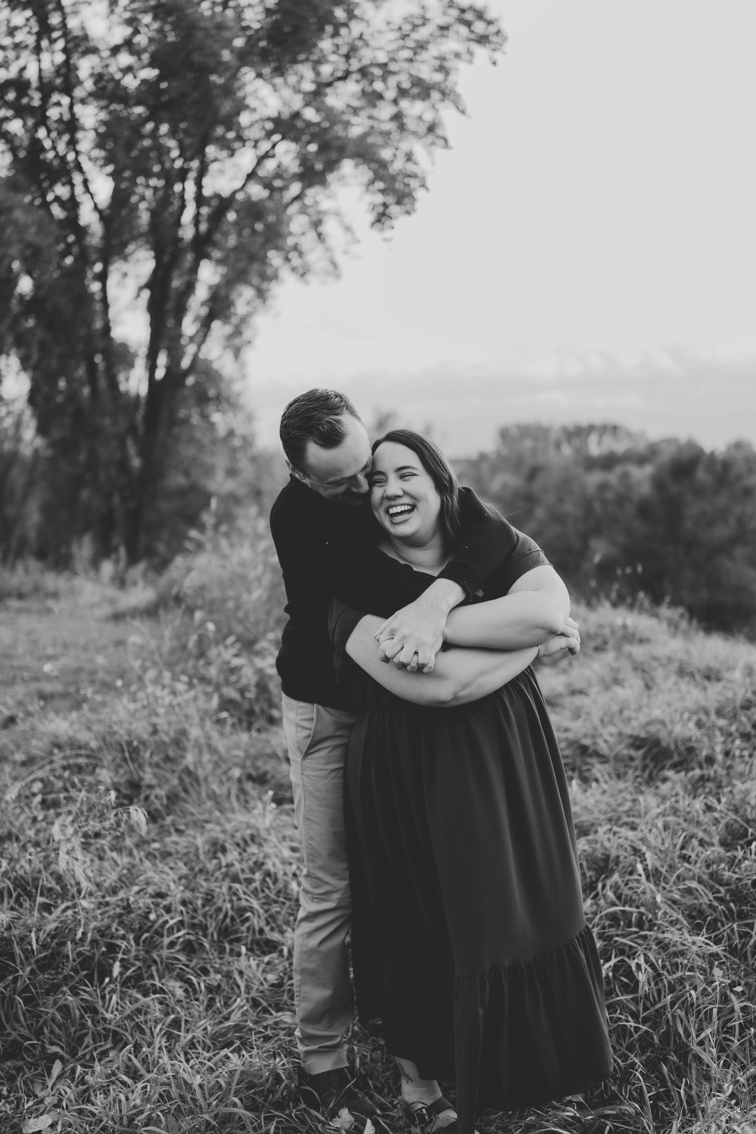 Black and white photo of a man hugging a laughing woman from behind in a lush outdoor setting.