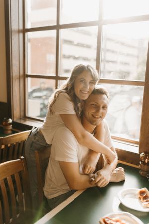 Smiling couple embracing by a sunlit restaurant window, woman hugging man from behind at a table.