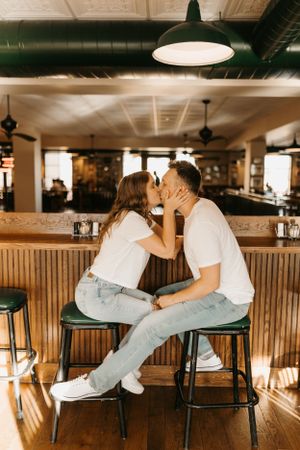 Couple sharing a romantic kiss while seated on bar stools inside a warmly lit restaurant.