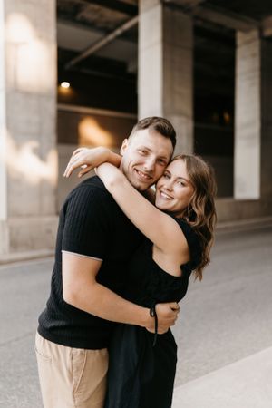 Happy couple hugging and smiling outdoors near concrete pillars, wearing matching black outfits.