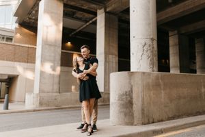 Couple in black outfits embracing and laughing together on a city sidewalk near stone columns.