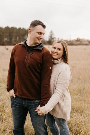 Couple holding hands in a golden autumn field, woman in cream sweater smiling at camera, man in burgundy pullover.