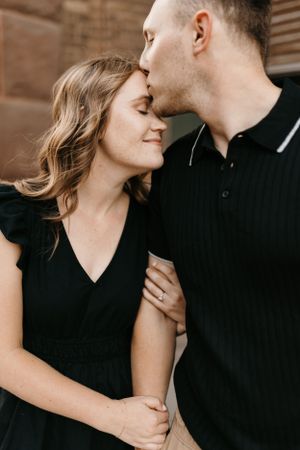Man kissing woman on forehead while holding hands, woman wearing black dress with engagement ring.
