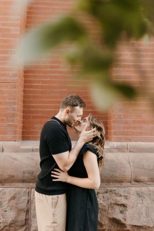 Romantic couple nearly kissing in front of a red brick wall, man in black shirt embracing woman.