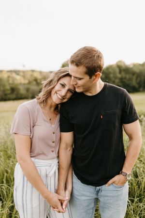 Couple embracing in a green field, woman smiling as man kisses her head during engagement session.
