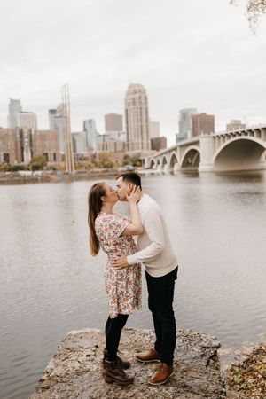 Couple kissing on rocky riverbank with Minneapolis skyline and bridge in the background.