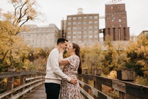 Couple sharing a kiss on a wooden bridge with autumn trees and brick building in background.