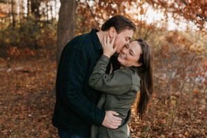 Couple laughing and embracing in autumn woods, man kissing womans cheek among colorful fall foliage.