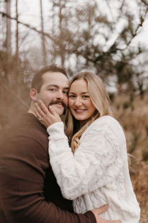 Smiling blonde woman in white knit sweater embracing man in brown sweater outdoors in wooded setting.