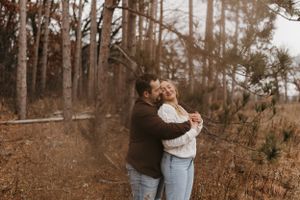 Couple laughing and embracing among tall pine trees in autumn forest, woman in white sweater and jeans.
