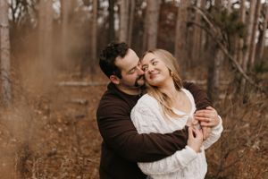 Man in dark brown sweater kissing smiling woman in white knit sweater in autumn woodland setting.