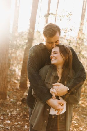 Couple sharing a tender embrace in sunlit forest, man hugging woman from behind among golden trees.