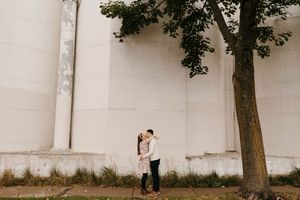 Couple kissing beside a large tree against white industrial silos during outdoor engagement session.