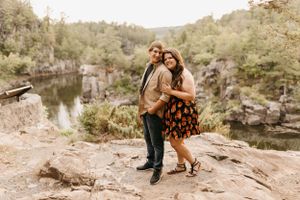 Smiling couple posing on rocky overlook above a scenic quarry lake surrounded by lush forest.