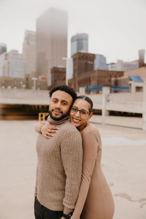 Couple embracing on city street, woman in tan dress hugging man in brown knit sweater with skyline behind.