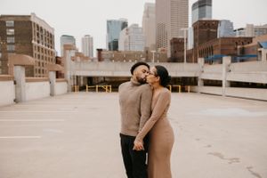 Couple sharing a kiss on rooftop parking lot with city skyline in background, dressed in coordinating earth tones.