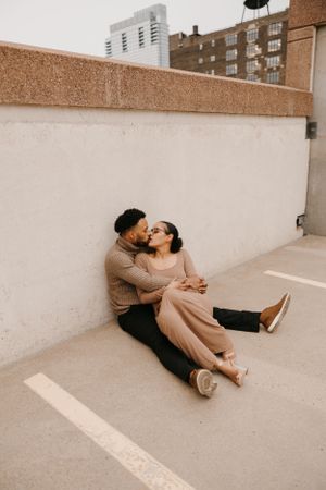 Couple sitting against wall on rooftop, sharing a kiss in matching earth-tone outfits with city buildings behind.