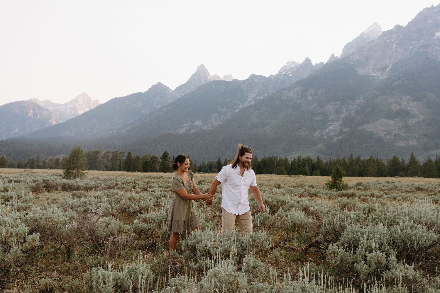 A couple holds hands in a wildflower field with majestic Grand Teton mountains rising in the background at dusk.