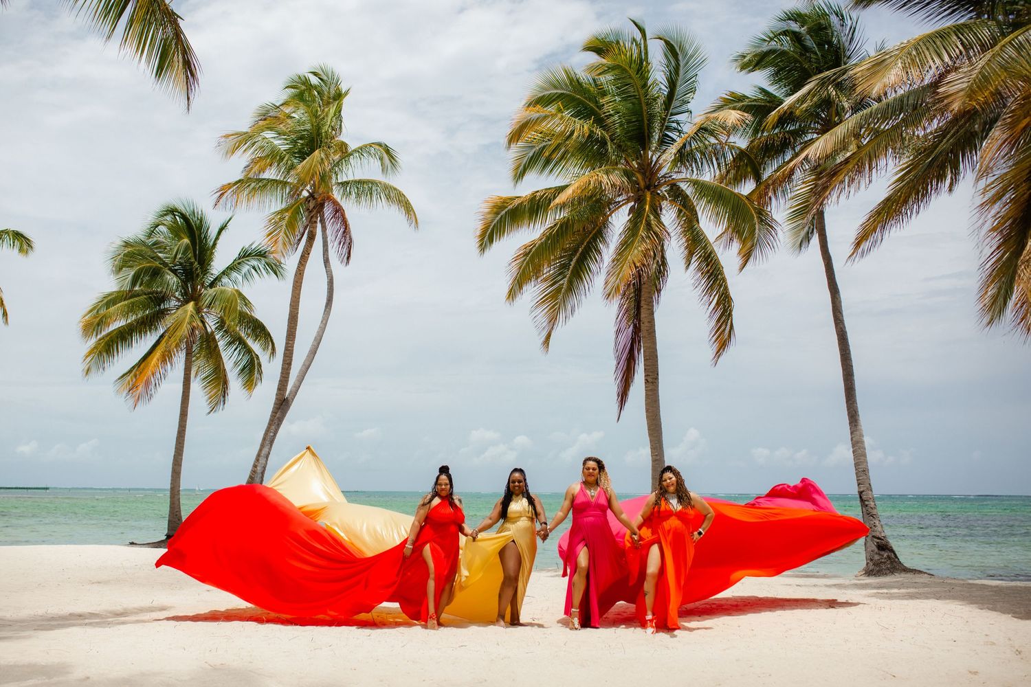 Palm trees sway in the wind along a tropical beach as people in red dresses pose for photos at sunset.