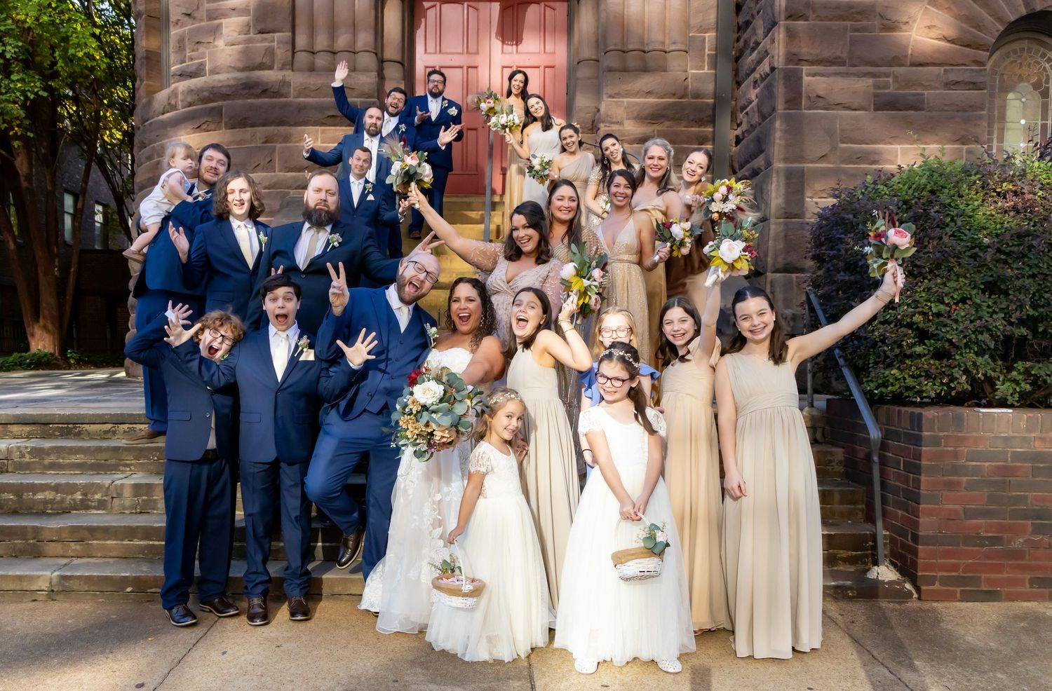 Wedding portrait at Wedding party in navy suits and cream dresses pose joyfully on church steps with flowers and decorative details.