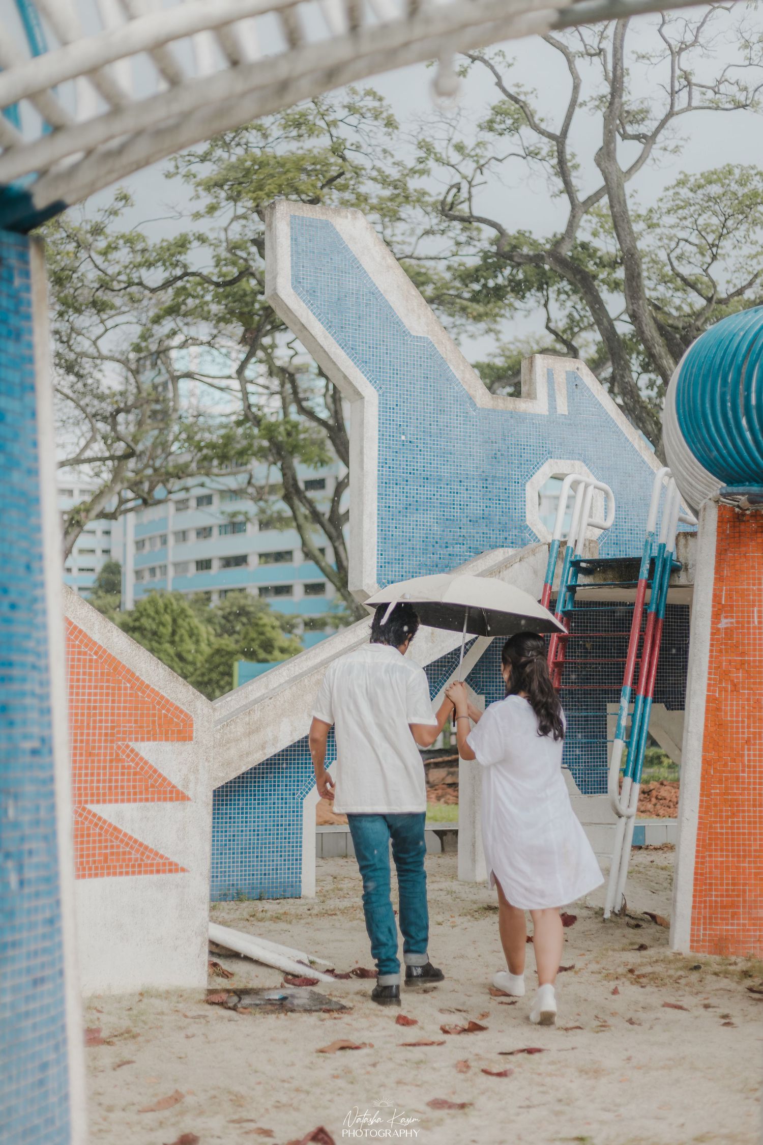 Couple sharing umbrella at colorful playground in Singapore, walking on sandy ground with blue mosaic structures.