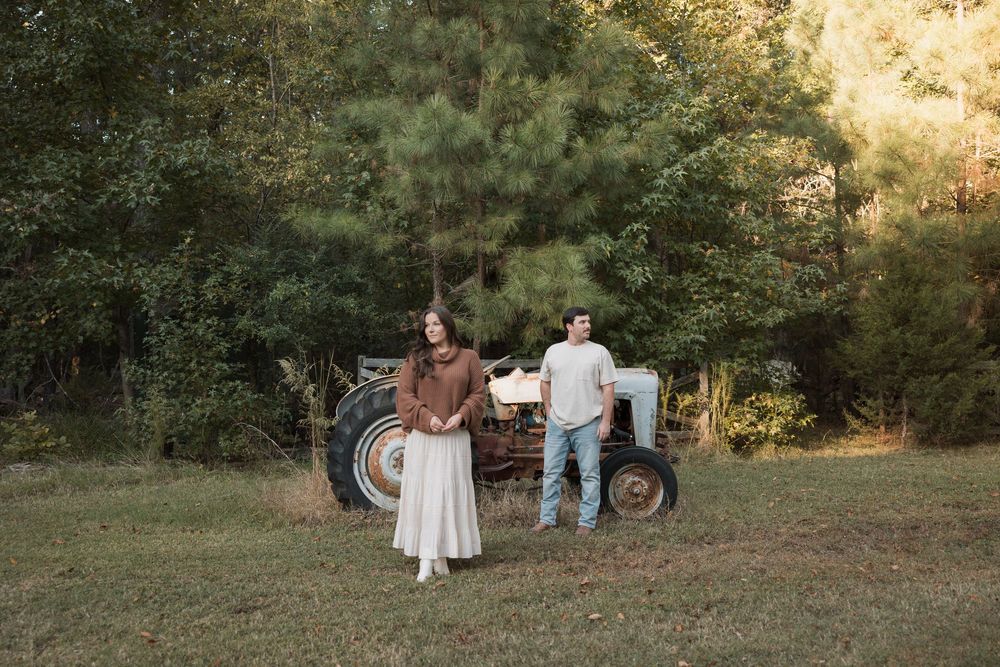 Couple posing in a rustic field with a vintage tractor and tall pine trees in the background.
