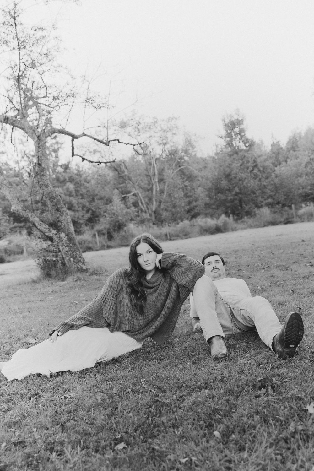 Black and white photo of a couple relaxing on grass in a park, woman in oversized sweater leaning against man.
