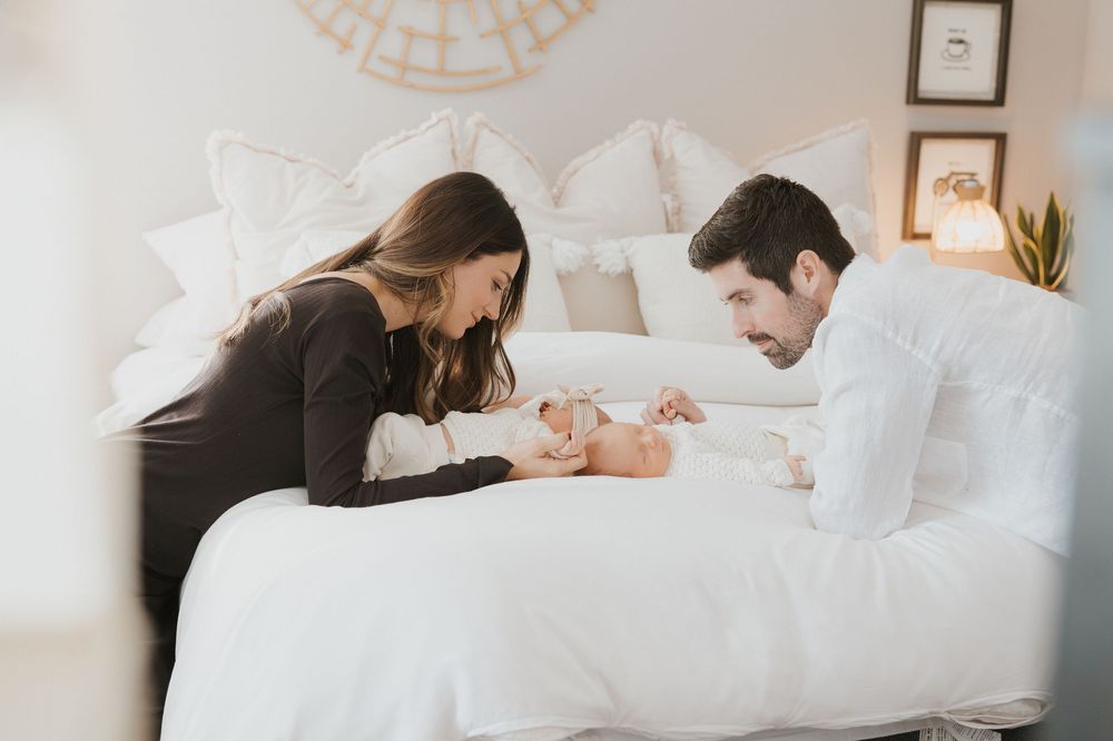 Parents lovingly gazing at their newborn baby while lying together on white bedding.