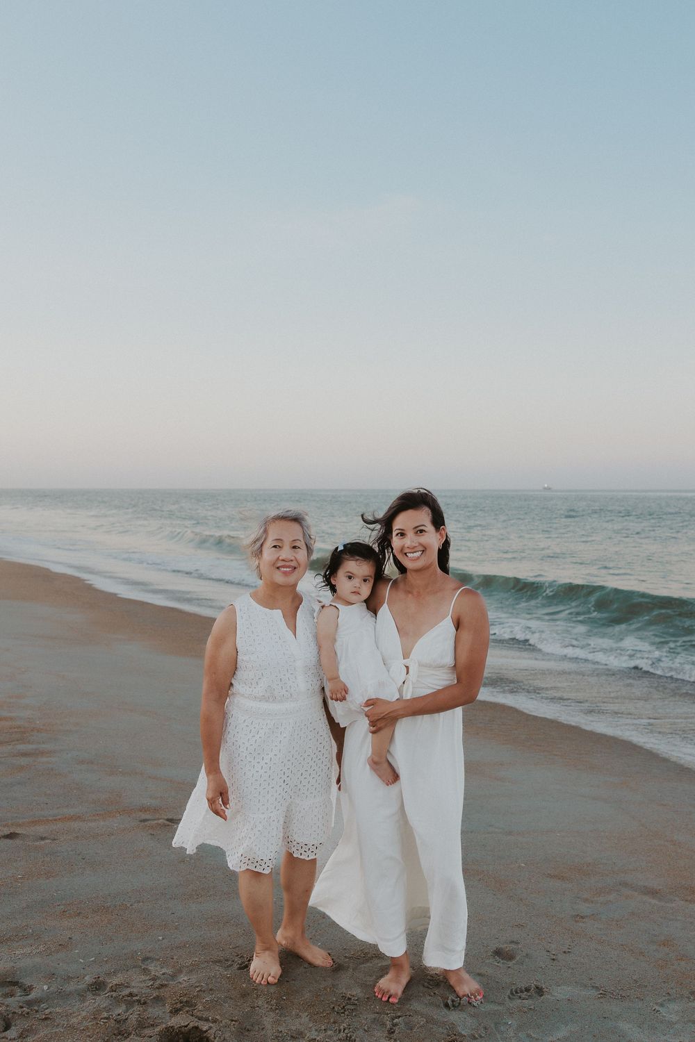 Family wearing white outfits walking together on a sandy beach at sunset with ocean waves in the background.