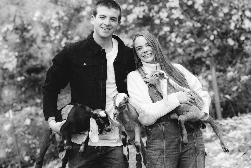 A smiling couple poses outdoors in black and white, each holding a small goat in their arms.