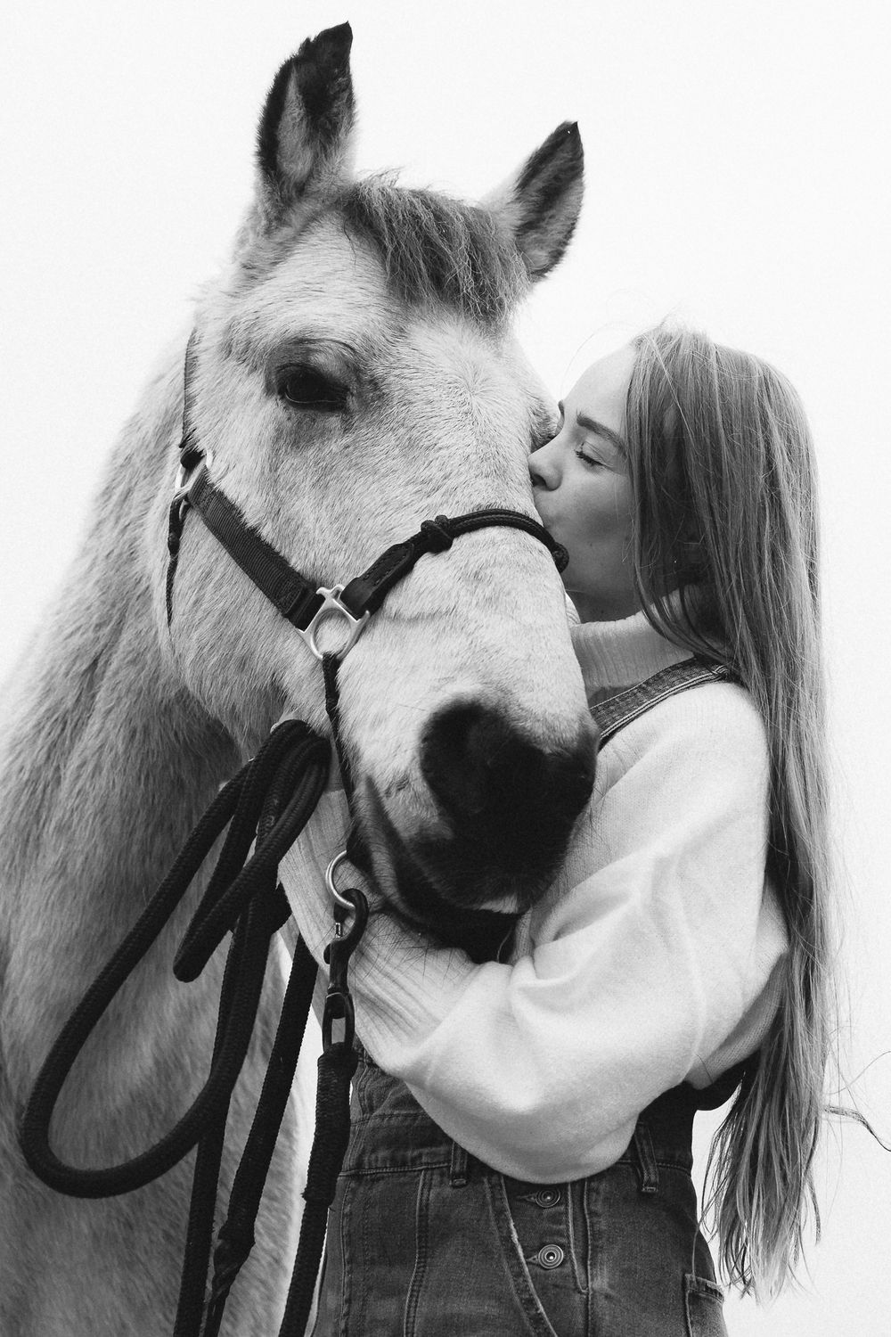A young woman with long hair tenderly kisses a white horse on the nose in a black and white photo.
