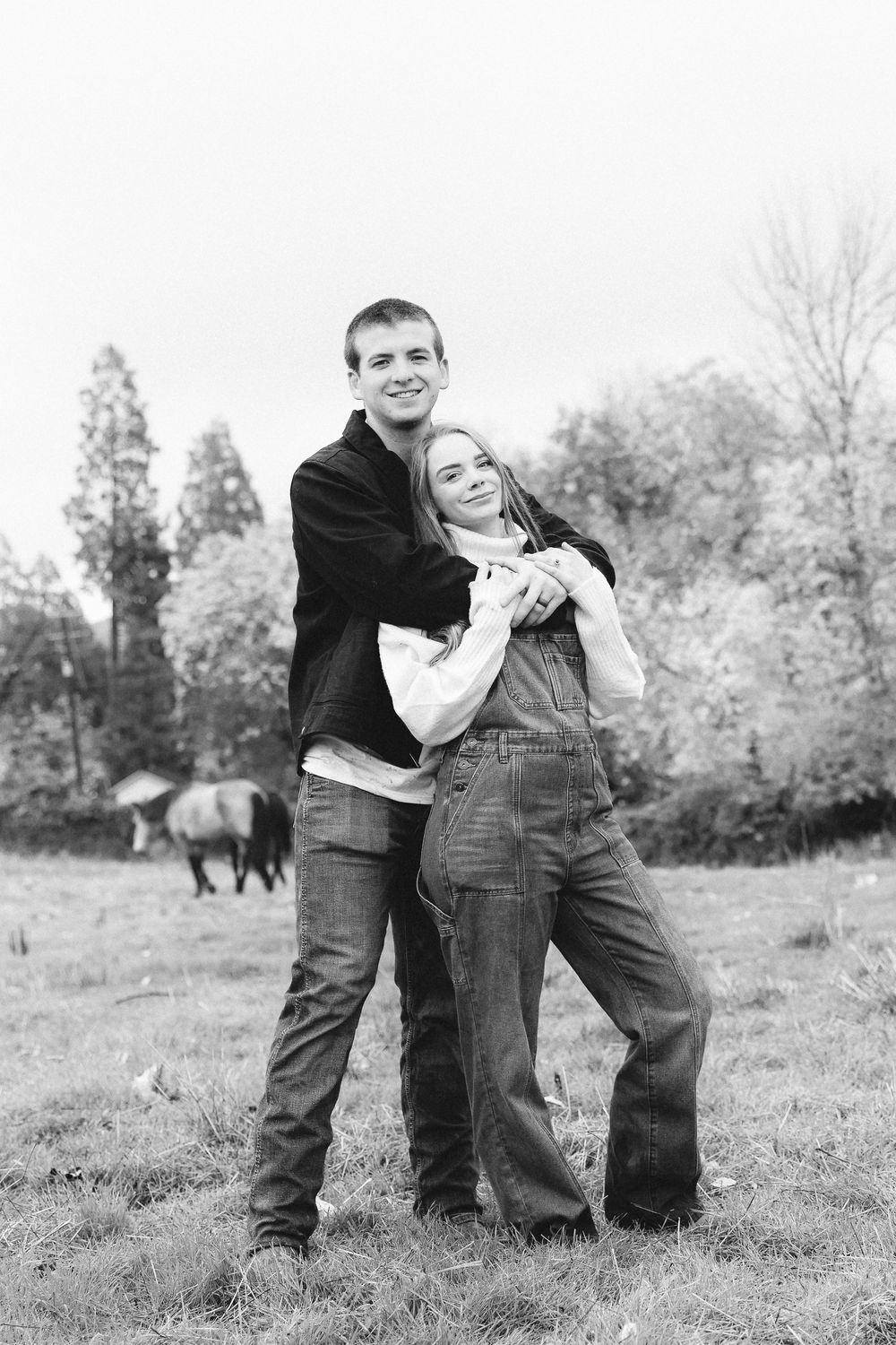 Black and white photo of a young couple embracing in a field with a horse and trees in the background.