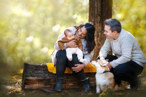 A happy family sits on a log in an autumn forest with their white fluffy dog while enjoying the golden afternoon light.