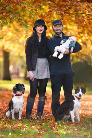 A couple stands with their baby and two border collies under vibrant autumn foliage in a park setting.