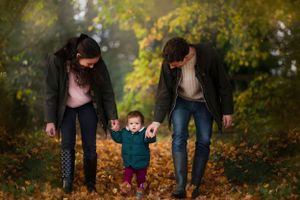 A young child in a red sweater walks hand in hand with parents on a path covered with autumn leaves.