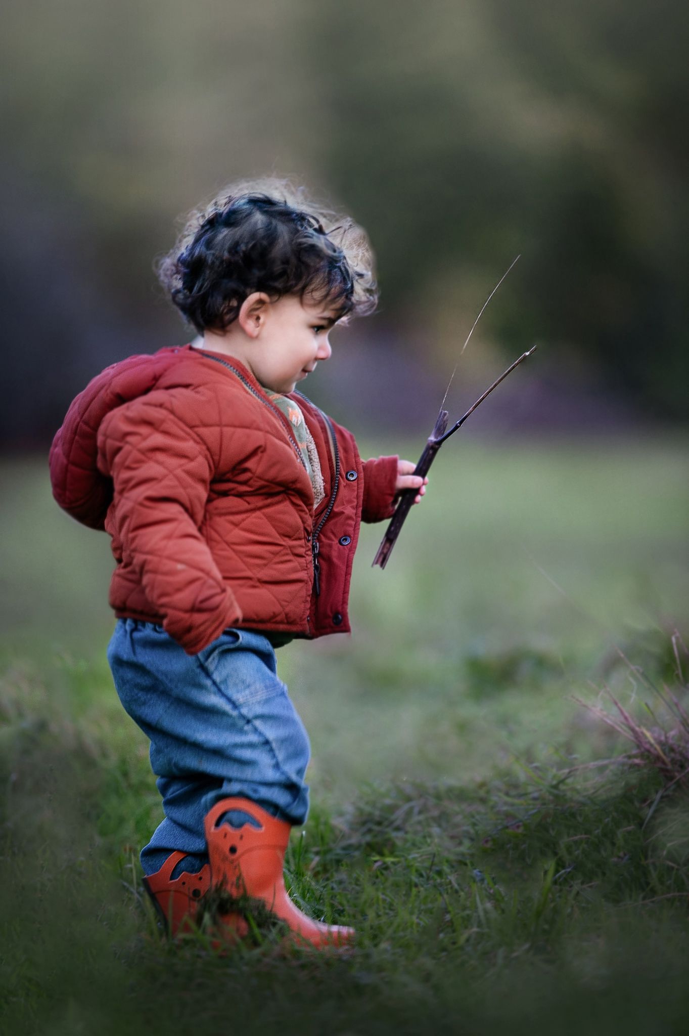 A small child in a red winter coat and jeans stands in tall grass while holding a fishing pole.