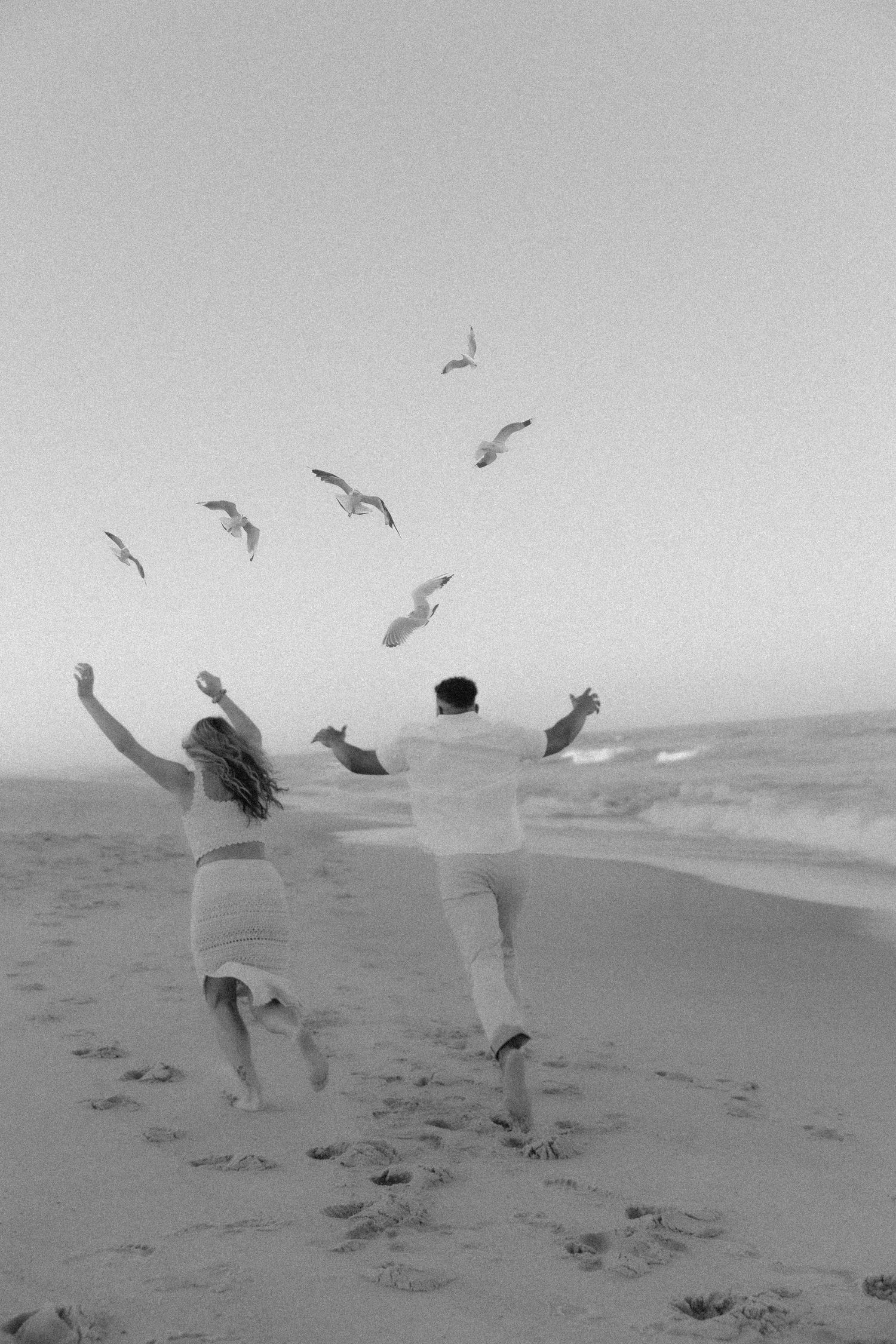 Two people joyfully leap on a beach as seagulls soar overhead in a black and white photograph.