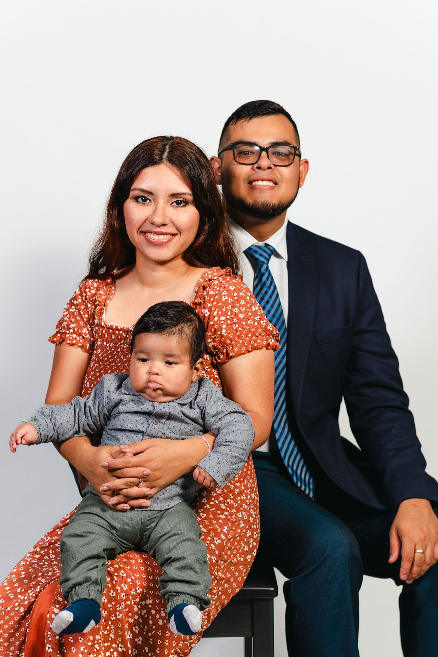 Family portrait with smiling parents and baby in formal attire against white background.