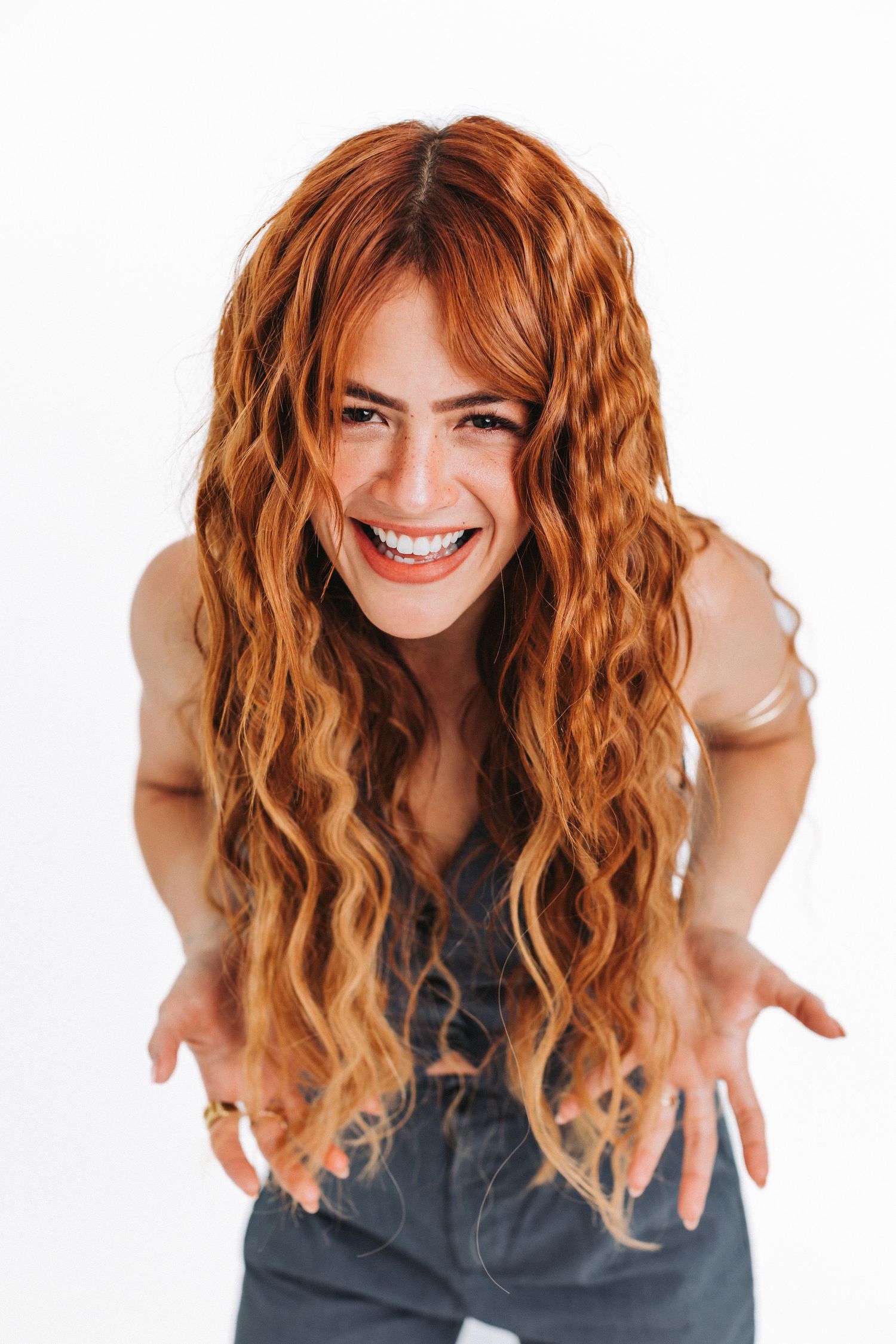 Woman with long wavy copper hair smiling while leaning forward in dark top against white background.