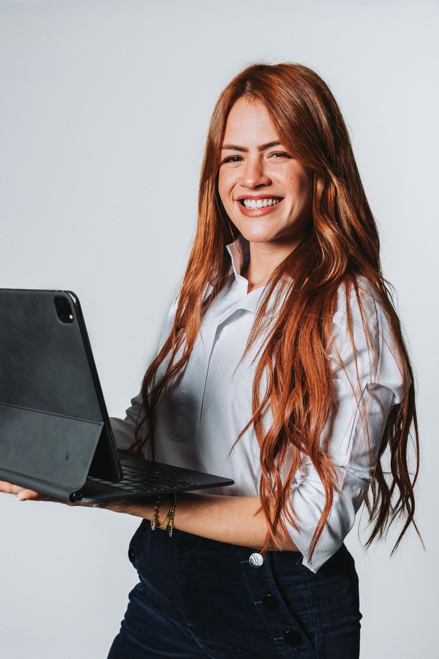 Woman in business attire smiling while holding a laptop against white background.