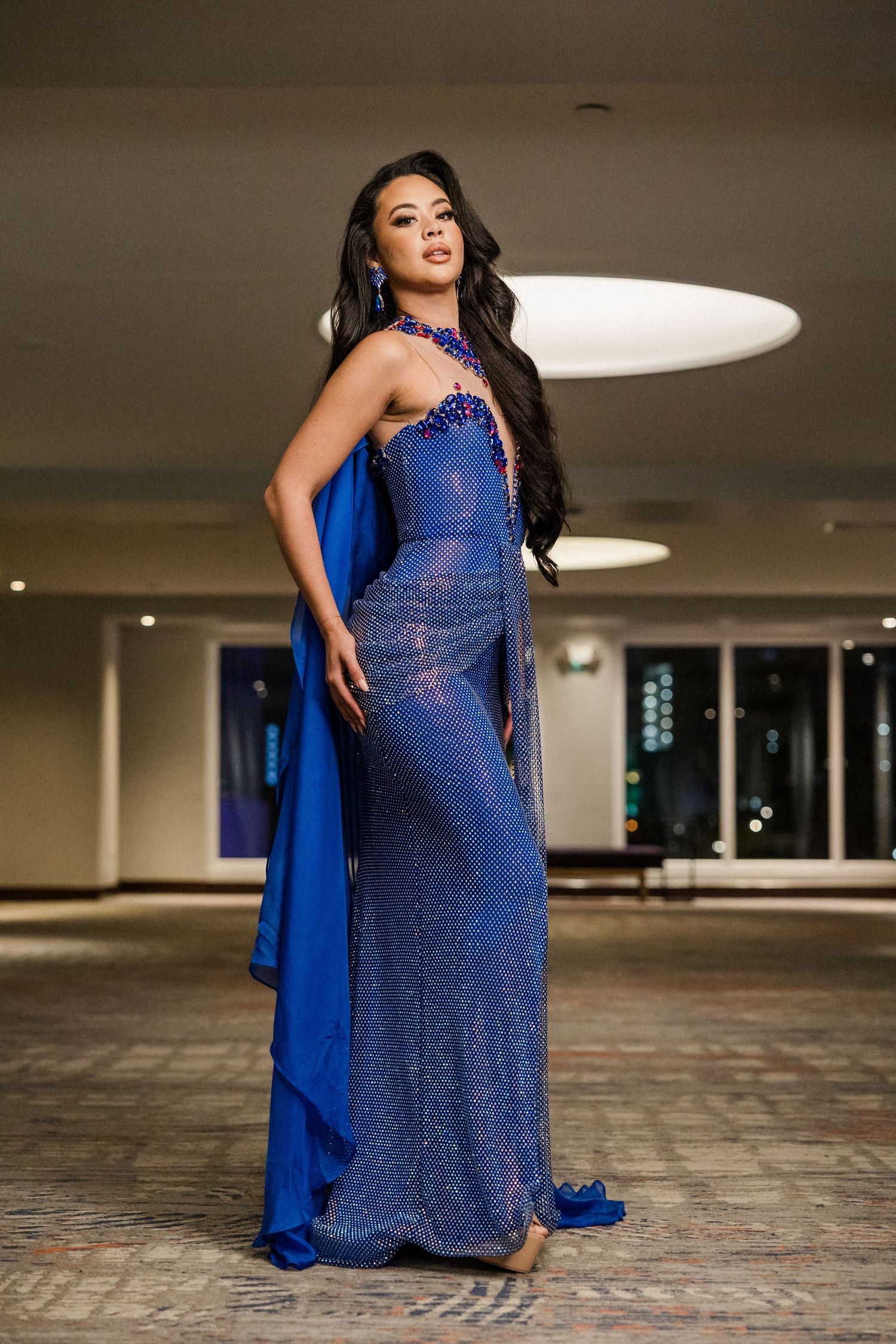 Elegant evening gown in royal blue sparkles with a flowing cape detail and fitted silhouette against urban backdrop.