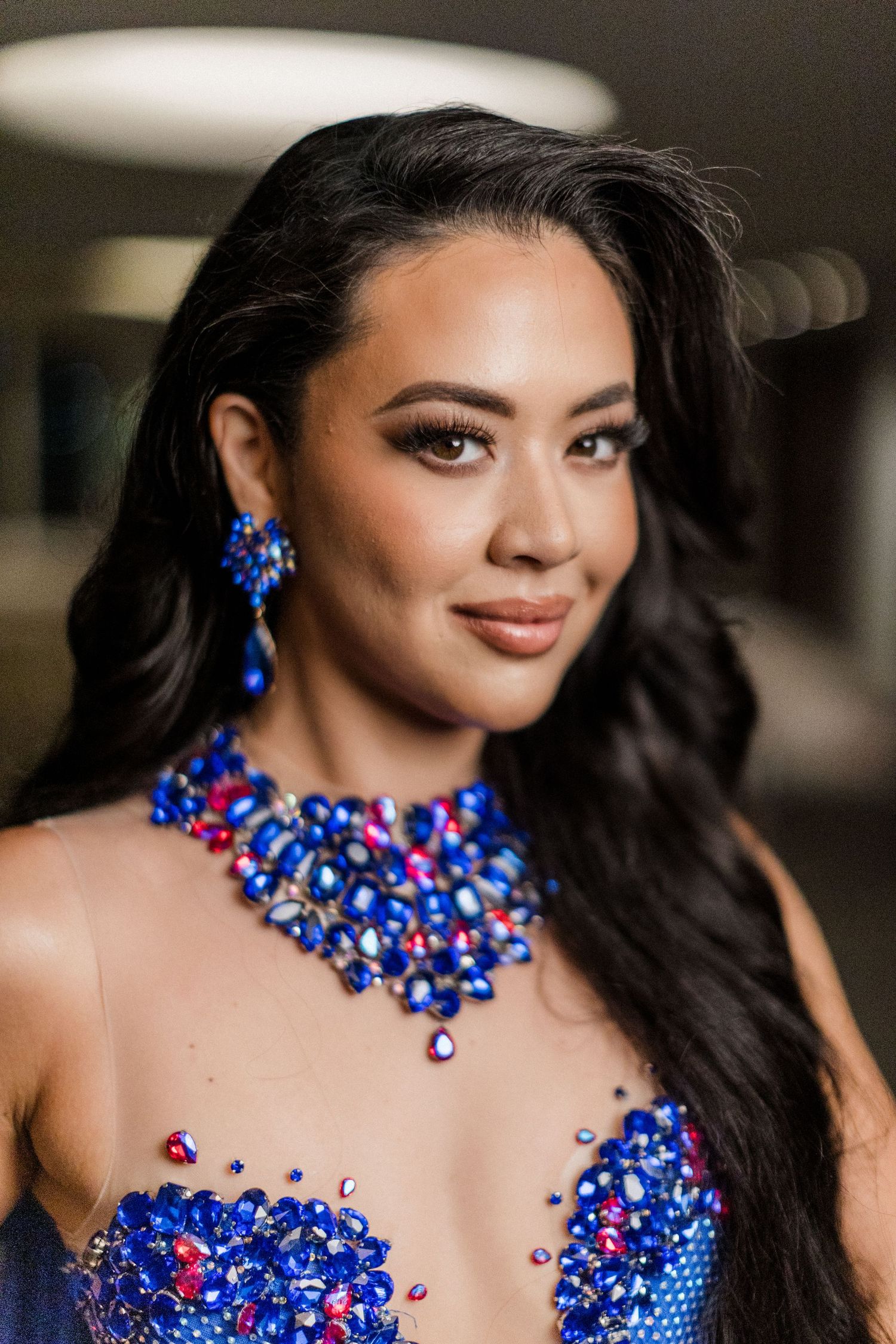 Close-up portrait showing wavy dark hair and glamorous statement jewelry in vibrant royal blue and red crystals.